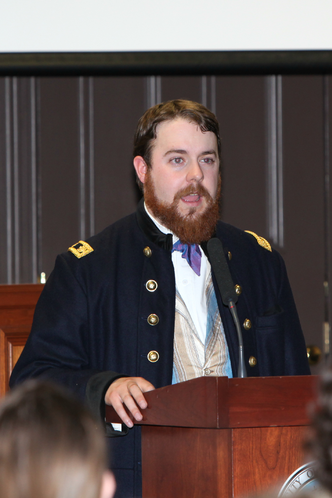 A young, bearded man in Civil War attire speaks at a microphone.