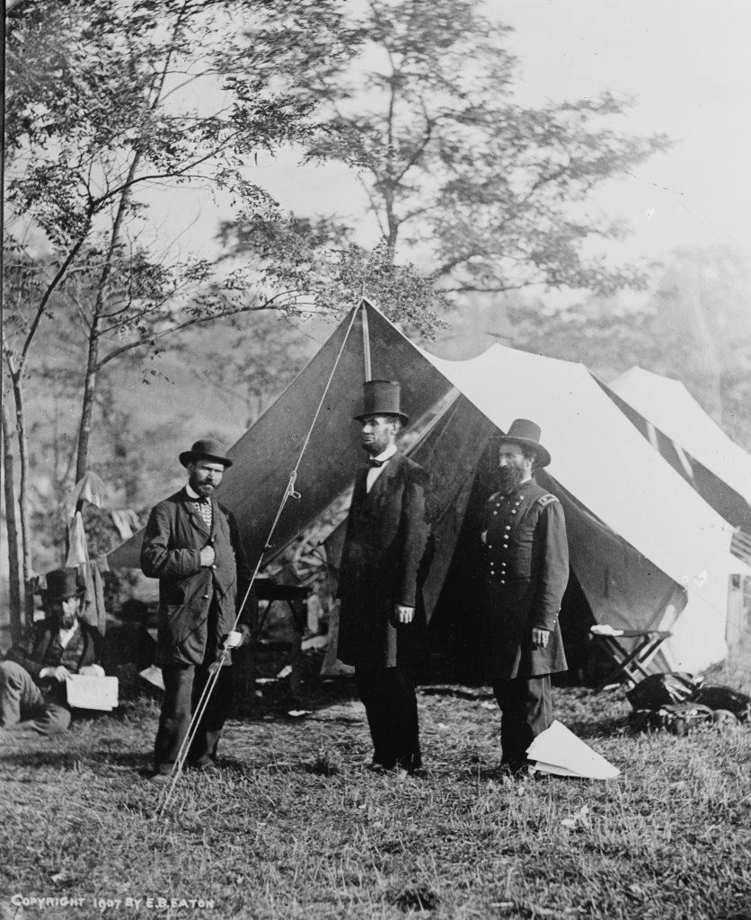 A historic black and white photograph of President Lincoln and two other men below a tree on a battlefield.